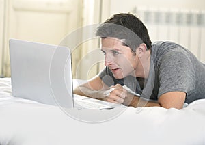 Young attractive man lying on bed or couch enjoying social networking using computer laptop at home