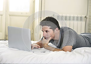 Young attractive man lying on bed or couch enjoying social networking using computer laptop at home wireless internet