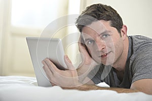 Young attractive man lying on bed or couch enjoying social networking using digital tablet computer internet at home