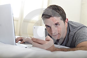 Young attractive man lying on bed or couch using mobile phone and computer laptop internet addict