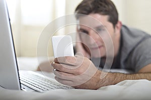 Young attractive man lying on bed or couch using mobile phone and computer laptop internet addict