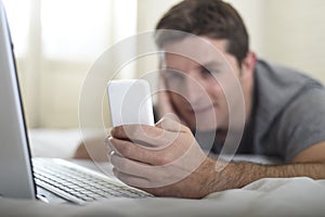 Young attractive man lying on bed or couch using mobile phone and computer laptop internet addict