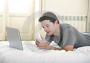 Young attractive man lying on bed or couch using mobile phone and computer laptop working from home