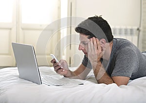 Young attractive man lying on bed or couch using mobile phone and computer laptop working from home