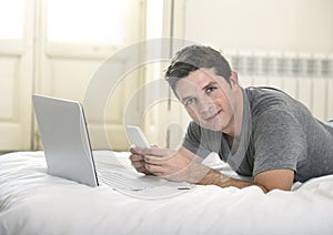 Young attractive man lying on bed or couch using mobile phone and computer laptop working from home