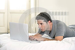 Young attractive man lying on bed or couch working on computer laptop typing connected to internet