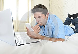 Young attractive man lying on bed using mobile phone and laptop working from home