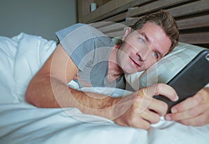 Young attractive man smiling happy lying on bed at night using social media app on mobile phone texting or having video call in co