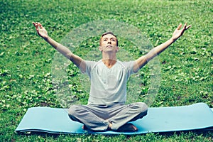 Young attractive man in sport clothes is meditating in the lotus position with a pacified face in the park against the background