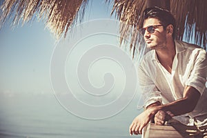 Young attractive man with sunglasses looking out over the sea during the summer. He looking forward, dressed in a white shirt and