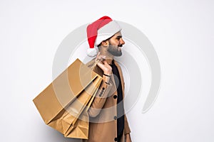 Young attractive man wearing Santa hat holding and pointing red shopping bag in christmas consumerism , December sale, buying xmas