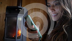 Young Attractive Mixed Race Woman Smiling and Using Mobile Phone near Fireplace. Cozy and Technology HD Slowmotion