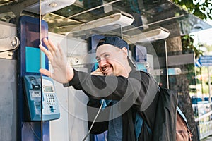 A young attractive modern man speaks at a payphone and welcomes with his hand. Communication concept.