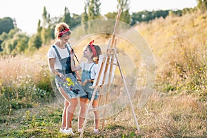 Young attractive mother teaches daughter painting in summer park. Outdoors activity for school age children concept.