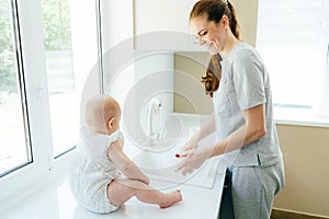 A young attractive mother washes the dishes in the sink while the baby is sitting on the table.