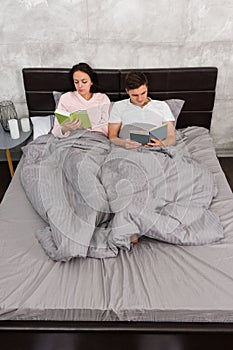 Young attractive pair reading books while lying in the bed and w