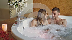 Young attractive people are hugging and talking in bubbling bathtub with foam in modern day spa. Romantic relationship