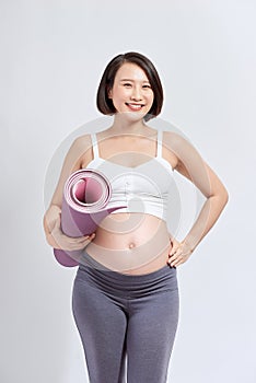 Young attractive pregnant female touching her belly while holding yoga mat in hands, expectant mother doing physical exercises,