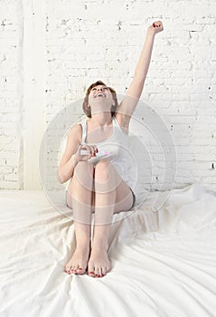 Young attractive pregnant woman holding happy positive result pregnancy test sitting on bed