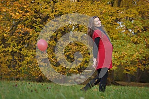 Young attractive pregnant woman in red dress, walking with small red balloon in hands and smiling. Autumn park