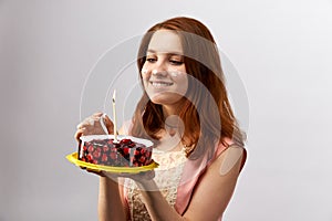 Young attractive red-haired girl holding a cake with candle and makes a wish on the birthday.