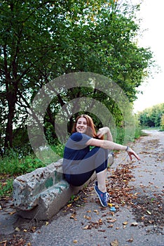 A young attractive red-haired girl is sitting on her haunches leaning against an old sidewalk stone.