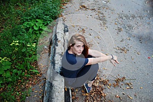 A young attractive red-haired girl is sitting on her haunches leaning against an old sidewalk stone. The girl smiles sweetly.