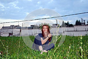 A young attractive red-haired girl with a wreath of wild flowers on her head is sitting on the grass on a warm summer day.