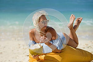 Young attractive and relaxed blond woman in bikini lying on beanbag hammock at tropical paradise beach drinking coconut water