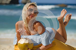 Young attractive and relaxed blond woman in bikini lying on beanbag hammock at tropical paradise beach drinking coconut water