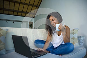 Young attractive and relaxed latin American student woman sitting at home sofa couch networking with laptop computer drinking coff