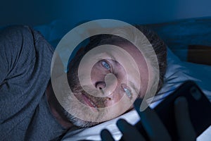 Young attractive and relaxed man with blue eyes lying on bed late at night in dark and dim light networking on mobile phone or