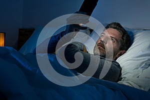 Young attractive and relaxed man with blue eyes lying on bed late at night in dark and dim light networking on mobile phone or