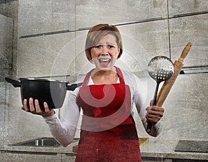 Young attractive rookie home cook woman in red apron at home kitchen holding cooking pan and rolling pin screaming desperate