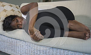 Young attractive and sad black African American woman lying in fetal position depressed at home sofa couch suffering menstruation