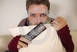 Young attractive scared and stressed man at living room couch holding TV remote watching horror movie or scary news on television