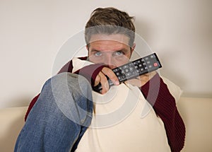 Young attractive scared and stressed man at living room couch holding TV remote watching horror movie or scary news on television