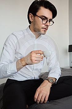 Young attractive serious brunette man in white shirt and eyeglasses sitting on bed with cup of coffee in hand