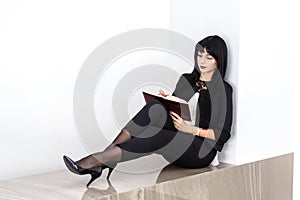 Young Attractive serious brunette woman dressed in a black business suit sitting on a floor in a office, writing in a notebook