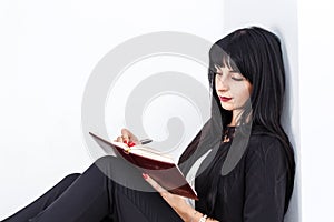 Young Attractive serious brunette woman dressed in a black business suit sitting on a floor in a office, writing in a notebook