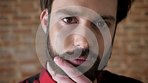 Young attractive serious man is touching his beard, watching at camera, brick background