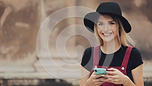 Young Attractive Smiling Blond Girl With Nose Jewelry Wearing a Black Hat Using her Smartphone Typing and Looking to