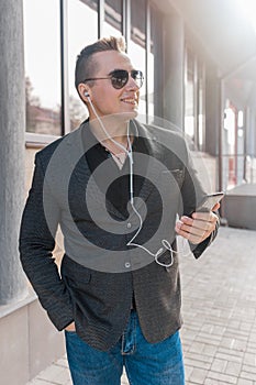 A young attractive smiling businessman of European appearance in a jacket and shirt style, wears sunglasses and listens to music