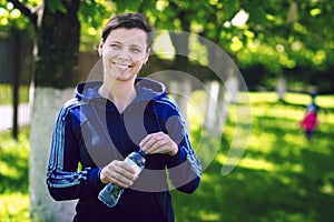 Young attractive smiling girl in sports sweatshirt with bottle of water in her hand in green summer park