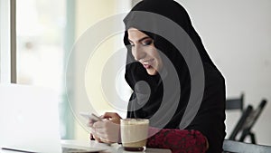 Young attractive smiling muslim woman in hijab sitting in the cafe and typing a message on her mobile phone. Laptop and