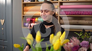 Young attractive smiling woman in apron working in floral shop and arranging bunch of flower using fresh yellow tulips
