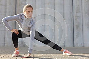 Young attractive sporty girl warming up outdoors, doing stretching at sunset or sunrise in city