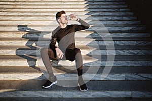 Young attractive sporty man sitting on stairs while drinking water after workout outdoor