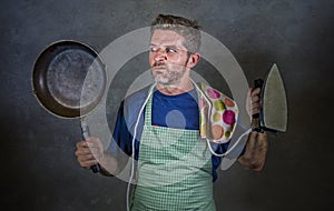 Young attractive stressed and overwhelmed lazy man holding kitchen pan and iron in stress and frustrated face expression