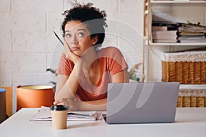 Young attractive student girl with dark curly hair sitting at the table with laptop and cup of coffee to go leaning on